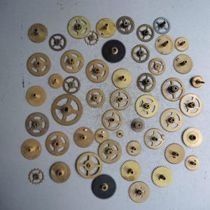 Op de afbeelding: Een verzameling messing uurwerktandwielen van verschillende groottes, verspreid over een grijze ondergrond. De tandwielen variëren in grootte en ontwerp, sommige hebben meerdere spaken en andere een massief midden. Er zijn ook een paar zwarte tandwielen aanwezig.
