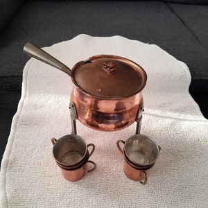 May include: A set of copper mugs and a copper pot with a lid and a handle. The pot is on a stand with three legs. The mugs are stacked on top of each other.