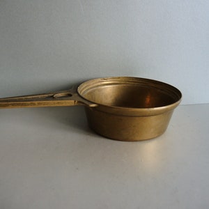 Puede incluir: Una taza medidora de latón con asa. La taza tiene una forma redondeada y una superficie lisa.