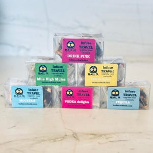 May include: A collection of clear, square boxes, each containing cocktail infusion kits. Each box has a colorful label with the text "infuse TRAVEL" and a flavor name like "DRINK PINK" or "TEQUILA". The brand name "HAIL M" is also visible.