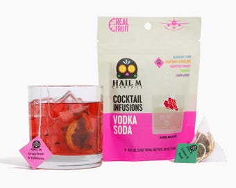 May include: A cocktail in a clear glass with ice and a pink infusion tea bag. A package of cocktail infusions with the text "Vodka Soda" and "Hail M Cocktails" is next to the glass. The package is white and pink.
