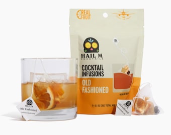 May include: A clear glass of an Old Fashioned cocktail with an orange slice and ice, next to a package of cocktail infusions. The package is beige with the text "HAIL M COCKTAILS COCKTAIL INFUSIONS OLD FASHIONED". A tea bag is in the glass.