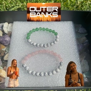 Könnte beinhalten: Zwei weiße und graue Perlenarmbänder mit einem grünen und weißen Perlenarmband darüber. Die Armbänder befinden sich in einer weißen Box mit dem Schriftzug "Outer Banks" auf einem Aufkleber.