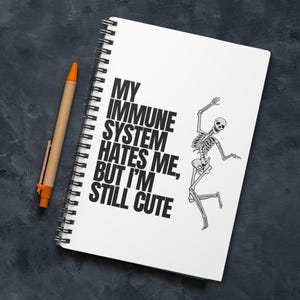 May include: A white spiral-bound notebook with the text "MY IMMUNE SYSTEM HATES ME, BUT I'M STILL CUTE" and a dancing skeleton illustration. A pen with a tan barrel and orange accents rests beside the notebook on a dark gray surface.