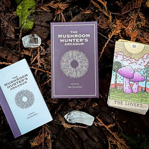 May include: Two purple books titled "The Mushroom Hunter's Arcanum" with a white circle design on the cover. A tarot card with a yellow sun and two purple mushrooms labeled "The Lovers" is on a bed of brown and green leaves.
