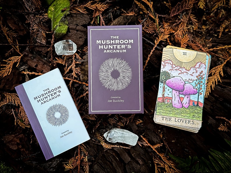 May include: Two purple books titled "The Mushroom Hunter's Arcanum" with a white circle design on the cover. A tarot card with a yellow sun and two purple mushrooms labeled "The Lovers" is on a bed of brown and green leaves.