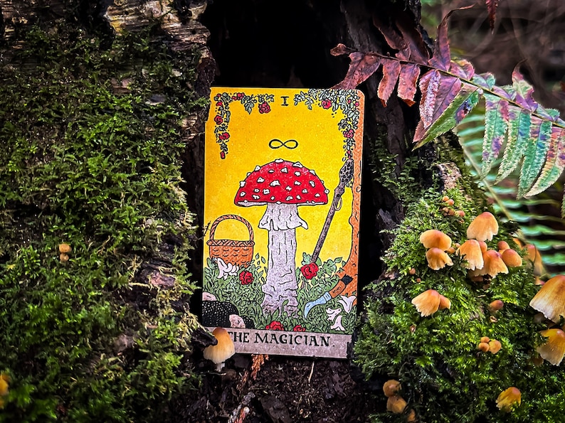 May include: A tarot card with a yellow background and a red and white mushroom in the center. The card is titled "The Magician" and has an infinity symbol above the mushroom. The card is surrounded by green foliage and a basket.