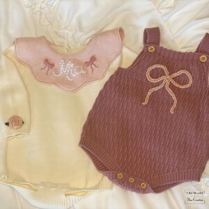 May include: Two knitted baby outfits, one white with a pink collar and the word "Mia" embroidered on it, and the other a maroon romper with a large brown bow.