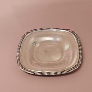 Pu&ograve; includere: Un piatto quadrato color argento con angoli arrotondati. Il bordo del piatto presenta un motivo dettagliato e scanalato. Il piatto &egrave; vuoto e riflette la luce. Lo sfondo &egrave; rosa tenue.