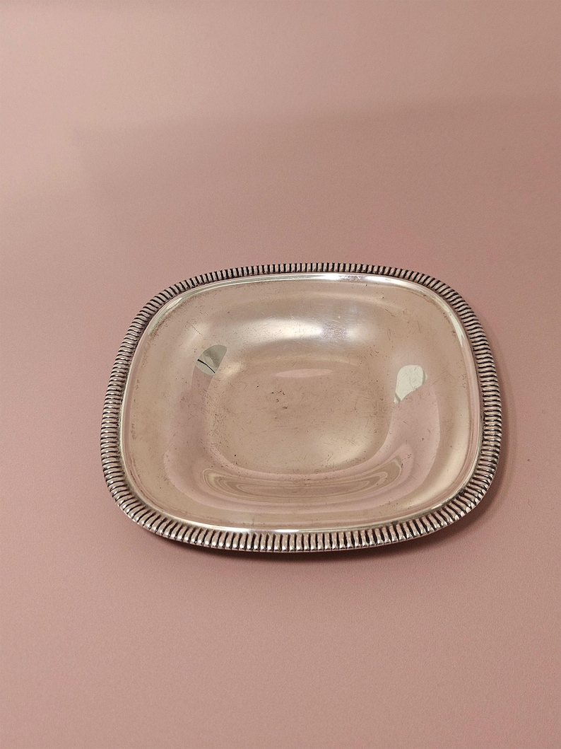 Pu&ograve; includere: Un piatto quadrato color argento con angoli arrotondati. Il bordo del piatto presenta un motivo dettagliato e scanalato. Il piatto &egrave; vuoto e riflette la luce. Lo sfondo &egrave; rosa tenue.
