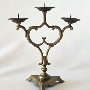 Könnte beinhalten: Ein Messing-Kandelaber mit drei Kerzenhaltern. Der Kandelaber hat ein verziertes, geschwungenes Design mit quadratischem Sockel. Die Kerzenhalter sind kreisförmig und halten die Kerzen aufrecht. Das Messing hat eine warme, antike Oberfläche.