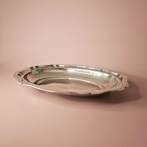 May include: A silver oval serving tray with a decorative, scalloped edge. The tray has a reflective surface and is set against a plain pink backdrop. The tray is empty and appears to be made of metal.