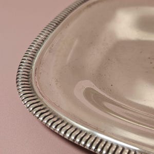 Pu&ograve; includere: Primo piano di un vassoio rettangolare color argento con angoli arrotondati. Il vassoio ha un bordo decorativo e scanalato. La superficie del vassoio &egrave; riflettente e mostra una leggera usura. Lo sfondo &egrave; rosa tenue.