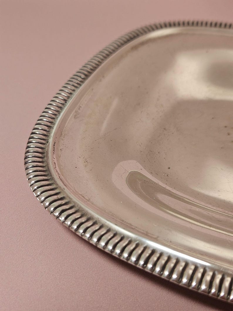 Pu&ograve; includere: Primo piano di un vassoio rettangolare color argento con angoli arrotondati. Il vassoio ha un bordo decorativo e scanalato. La superficie del vassoio &egrave; riflettente e mostra una leggera usura. Lo sfondo &egrave; rosa tenue.