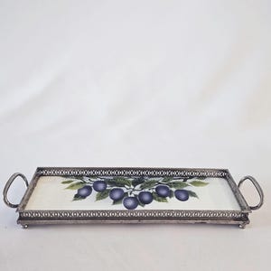 Può includere: Vassoio rettangolare con cornice e manici in metallo color argento. La superficie del vassoio presenta uno sfondo bianco con un disegno dipinto di prugne viola su un ramo con foglie verdi. Il vassoio è lungo circa 40 cm.