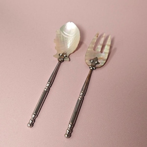 Può includere: Un set composto da cucchiaio e forchetta in metallo argentato con teste in madreperla iridescente. La testa del cucchiaio ha la forma di un pesce con dettagli a scaglie incisi, mentre la forchetta ha tre rebbi. Entrambi gli utensili hanno manici sottili e scanalati.