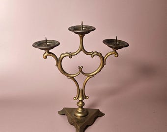 Può includere: Un candelabro in ottone con tre portacandele. Il candelabro ha un design ornato con una base triangolare. I portacandele sono circolari e hanno una piccola punta al centro per tenere le candele. Il candelabro è di colore oro.