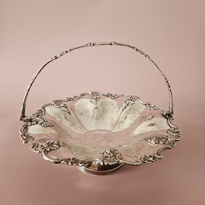 May include: Silver-toned decorative basket with a handle. The basket has a scalloped edge with floral detailing and a circular base. The interior of the basket has a floral pattern. The handle is arched and also has floral detailing.
