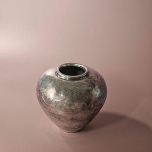 Puede incluir: Un jarrón de cerámica redondeado con una abertura circular estrecha y un acabado metálico. La superficie presenta una textura moteada con tonos mezclados de plata, gris y azul apagado, creando un aspecto reflectante en el cuerpo curvo.