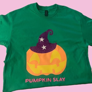 May include: Green t-shirt with a pumpkin wearing a purple witch hat with white stars. The text "PUMPKIN SLAY" is printed in pink below the pumpkin.