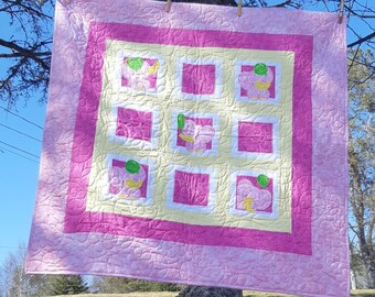 Puede incluir: Un edredón hecho a mano en tonos rosa, amarillo y blanco, colgado al aire libre. El edredón presenta un patrón repetitivo de cuadrados con diseños de patos bordados. El edredón está bordeado con un patrón floral rosa.