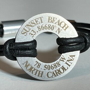 Puede incluir: Un brazalete de cuero negro con un dije de metal plateado. El dije está grabado con las palabras "Sunset Beach" y las coordenadas "33.86680° N 78.50688° W NORTH CAROLINA".