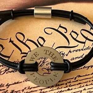 May include: A black leather bracelet with a silver metal charm that reads "We The People".