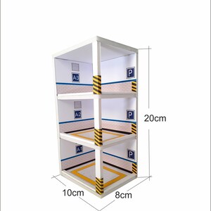 May include: A white cardboard model of a three-level parking garage with yellow and black striped barriers. The garage is labeled with the letters "A-1", "A-2", "A-3", and "P" for parking. The model measures 10 inches wide, 8 inches deep, and 20 inches tall.