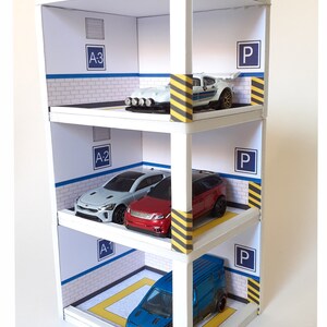 May include: A white cardboard multi-level parking garage with yellow and black striped barriers. The garage has three levels with a blue and white brick pattern and the letters "A-1", "A-2", and "A-3" on each level. The garage is filled with toy cars, including a yellow car on the top level, a white car on the second level, a red car and a silver car on the third level, and a blue car on the bottom level.