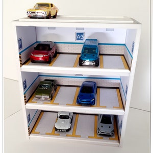 May include: A white wooden multi-level parking garage with five toy cars inside. The cars are a yellow muscle car, a red SUV, a blue truck, a blue sedan, and a white sedan. The garage has a sign that says "A3" on the top level.