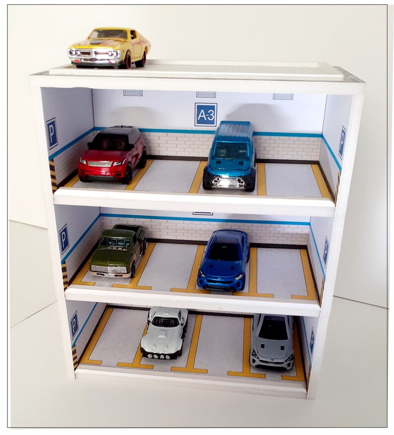May include: A white wooden multi-level parking garage with five toy cars inside. The cars are a yellow muscle car, a red SUV, a blue truck, a blue sedan, and a white sedan. The garage has a sign that says "A3" on the top level.