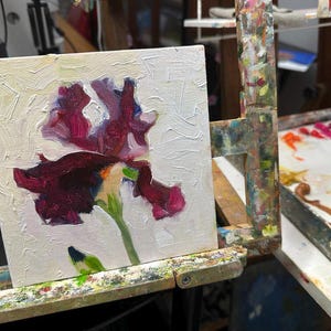 May include: An oil painting of a dark purple iris flower with green stem and leaves, set against a textured white background. The artwork is displayed on a wooden easel, showcasing the artist's work.