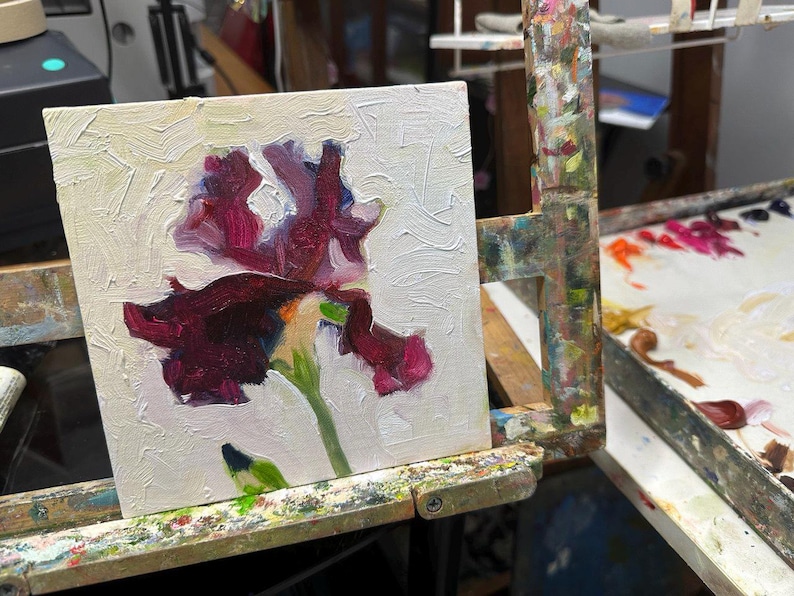 May include: An oil painting of a dark purple iris flower with green stem and leaves, set against a textured white background. The artwork is displayed on a wooden easel, showcasing the artist's work.