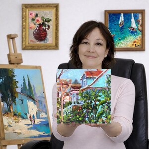The artist holding a square painting of a victorian house. Two other framed paintings are visible: one of roses in a vase and another of sailboats on the water. An easel holds a painting of a street scene.