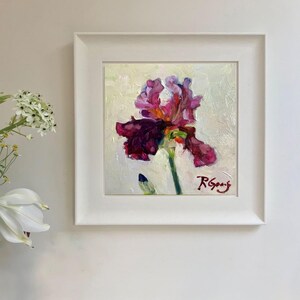 May include: Oil painting of a vibrant iris flower in shades of purple, magenta, and green, set against a light background. The artwork is framed in a white square frame, signed by the artist.