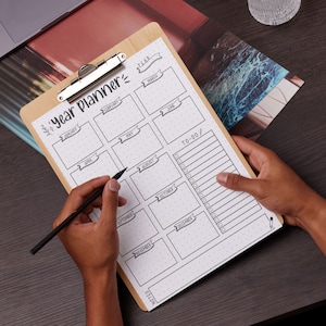 May include: A brown wooden clipboard with a year planner printed on it. The planner has a section for each month of the year, with a to-do list on the right side. A person is writing on the planner with a black pen.