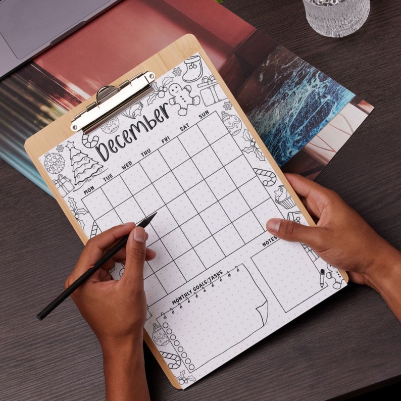 Puede incluir: Un portapapeles de madera con un dise&ntilde;o de calendario de diciembre. El calendario tiene una cuadr&iacute;cula para entradas diarias y una secci&oacute;n para objetivos mensuales. El dise&ntilde;o incluye garabatos festivos con temas navide&ntilde;os alrededor de los bordes.