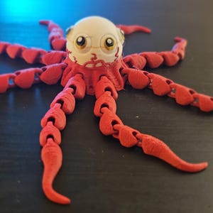 May include: A red articulated octopus toy with a white head and yellow-rimmed glasses. The octopus has eight segmented tentacles. The toy is made of plastic and sits on a dark surface.