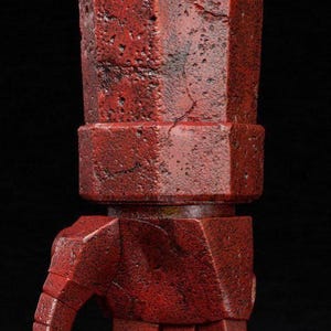 Puede incluir: Primer plano de una mano mecánica roja texturizada con un diseño geométrico. La mano está hecha de una serie de formas rectangulares y hexagonales, con una apariencia desgastada. El fondo es negro.