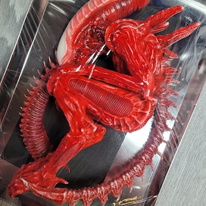 May include: A red alien creature figurine with a white egg sac. The figurine is curled up in a fetal position with its tail curled around its body. The figurine is made of plastic and has a detailed, realistic appearance.