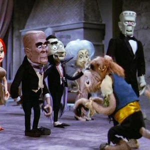 May include: A group of puppet characters, including a Frankenstein's monster, a vampire, and a red-haired woman, are gathered in a room. The puppets are dressed in formal attire, and the scene has a vintage, theatrical feel. The characters appear to be from a horror or monster-themed production.