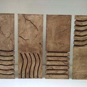 Puede incluir: Cuatro esculturas rectangulares marrones con superficies texturizadas y diseños tallados. Cada escultura tiene un patrón diferente, incluyendo líneas horizontales, líneas verticales y formas abstractas. Las esculturas están dispuestas una al lado de la otra.