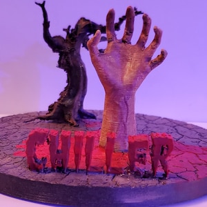 May include: A 3D printed horror themed figurine with a skeletal hand reaching up from a cracked ground base. The word "CHILLER" is printed in red on the base.