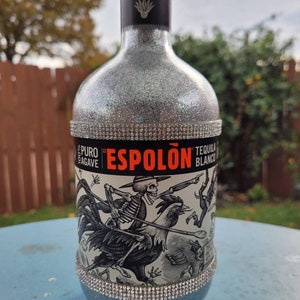 May include: A silver bottle of Espolón Tequila Blanco with a black and white illustration of a skeleton riding a rooster. The bottle is decorated with a silver glitter finish and a band of rhinestones around the middle.