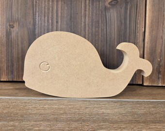 May include: A wooden whale-shaped cutout, featuring a smooth, light brown surface. The whale has a circular eye detail and a curved tail. The cutout is displayed against a wooden background.