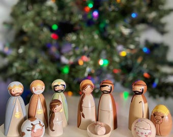 May include: A wooden nativity set with painted figures of Mary, Joseph, baby Jesus, the three wise men, a shepherd, a cow, a sheep, and a camel. The figures are all standing on a light brown surface in front of a blurred Christmas tree with lights.