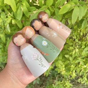 May include: Five wooden peg dolls with painted faces and brown hair. Each doll is wearing a different dress with a floral pattern in shades of green, brown, and pink.