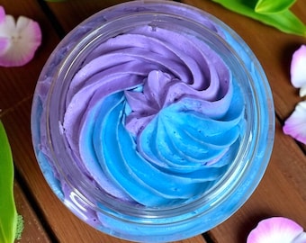 May include: Overhead shot of a clear jar filled with a swirl of blue and purple whipped cream. The jar is surrounded by green leaves and pink flower petals on a wooden surface. The cream has a smooth, textured appearance.