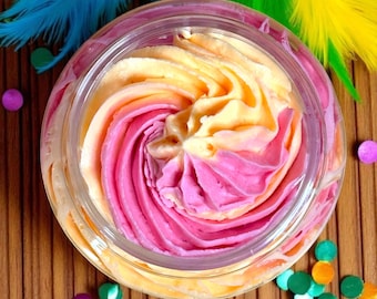 May include: A jar of colorful whipped body butter with a swirl design. The butter is a mix of pink and yellow, creating a vibrant contrast. The clear jar sits on a wooden surface, with colorful confetti and feathers in the background.