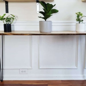 May include: A wooden console table with a live edge top and black hairpin legs. Three potted plants are on the table.